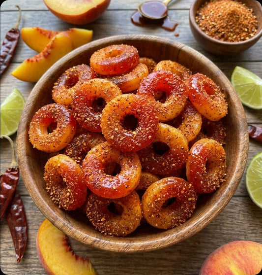 Wooden bowl filled with peach-flavored gummy rings coated in sugar and spice powder, surrounded by fresh peach slices, lime wedges, dried red chilies, and bowls of spice seasoning on a wooden surface