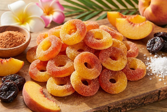 Wooden cutting board displaying peach-flavored gummy ring candies coated in sugar, surrounded by fresh peach slices, dried prunes, tropical plumeria flowers, palm leaves, cinnamon powder, and coarse sugar crystals